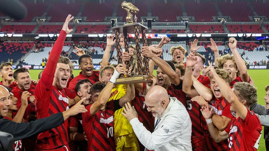 SIUE men's soccer team hoisting the Bronze Boot trophy in full celebration on the pitch at CITYPARK. It's nighttime and the field is brightly lighting their smiles.