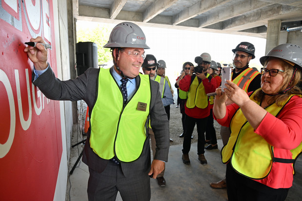 Dean Luer signs the wall and Dean Judy takes a photo of him doing that. Both are in hard hats.