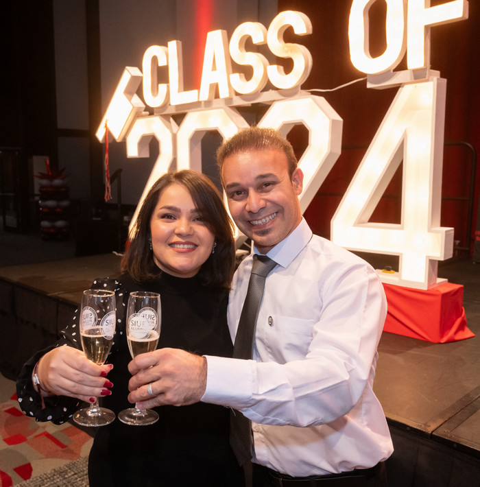 Two students cheering with champagne glasses in front of an illuminated Class of 2024 sign